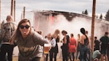 A group of people at an outdoor music festival with a stage and smoke effect in the background. A woman in the foreground playfully sticks her tongue out while holding a cup of beer, wearing sunglasses and casual attire.