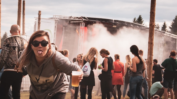 A group of people at an outdoor music festival with a stage and smoke effect in the background. A woman in the foreground playfully sticks her tongue out while holding a cup of beer, wearing sunglasses and casual attire.