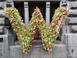 A large letter 'W' is formed entirely out of multicolored tulips. The arrangement is mounted on a stone building facade, with a narrow barred window in the background. The tulips are densely packed, creating a vibrant display of reds, yellows, pinks, and whites.