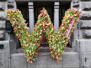 A large letter 'W' is formed entirely out of multicolored tulips. The arrangement is mounted on a stone building facade, with a narrow barred window in the background. The tulips are densely packed, creating a vibrant display of reds, yellows, pinks, and whites.