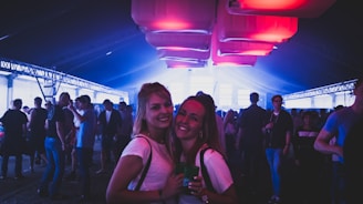 A lively scene of a crowded indoor event or party under soft purple and blue lighting. Two people in the foreground smiling at the camera, surrounded by groups of people engaged in conversations and activities. The ceiling features large glowing panels, adding a vibrant atmosphere.