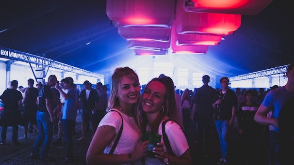 A lively scene of a crowded indoor event or party under soft purple and blue lighting. Two people in the foreground smiling at the camera, surrounded by groups of people engaged in conversations and activities. The ceiling features large glowing panels, adding a vibrant atmosphere.