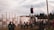 A diverse group of people gathers outdoors at a festival or event, featuring a stage with lighting and smoke effects. There are tall, wooden poles and an artistic canopy structure set against a backdrop of trees. The scene captures a relaxed and social atmosphere.