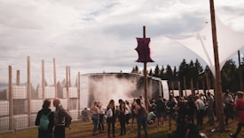 A diverse group of people gathers outdoors at a festival or event, featuring a stage with lighting and smoke effects. There are tall, wooden poles and an artistic canopy structure set against a backdrop of trees. The scene captures a relaxed and social atmosphere.