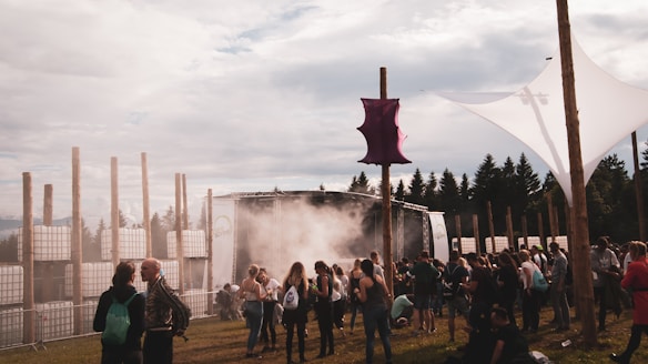 A diverse group of people gathers outdoors at a festival or event, featuring a stage with lighting and smoke effects. There are tall, wooden poles and an artistic canopy structure set against a backdrop of trees. The scene captures a relaxed and social atmosphere.