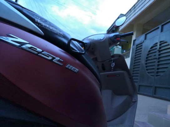A close-up view of a red scooter parked near a building. The scooter features a logo that reads 'Zest 110' in metallic letters on its body. The handlebars and mirrors are visible, and there is a key hanging from the ignition. In the background, part of a building and a gate can be seen, with a cloudy sky overhead.