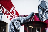 A large, expressive mural covers a wall, depicting a stylized figure with a focus on the hand pointing outward. The figure is rendered in geometric patterns, primarily using shades of gray, red, and white. The background features abstract shapes and letters, adding a dynamic element to the composition.