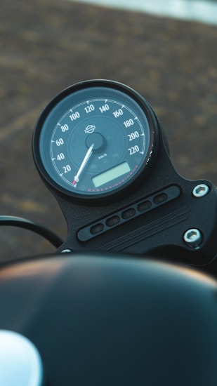 The image depicts a motorcycle speedometer with a circular dial. It displays speed measurements in kilometers per hour, ranging up to 220 km/h. The design features a minimalist aesthetic with clear markings and a small digital display at the bottom. The background appears out of focus, emphasizing the speedometer.