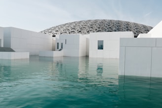 A modern architectural structure featuring white geometric buildings partially submerged in calm turquoise water. The background reveals a large, intricately patterned dome with a futuristic design.