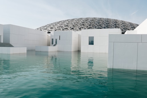 A modern architectural structure featuring white geometric buildings partially submerged in calm turquoise water. The background reveals a large, intricately patterned dome with a futuristic design.