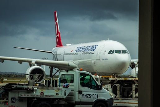 A large commercial airliner is parked on the tarmac with a service truck labeled 'Aircraft Lavatory Service' in the foreground. The airplane has a white body with a red tail fin featuring a logo. The sky is overcast, creating a moody atmosphere.