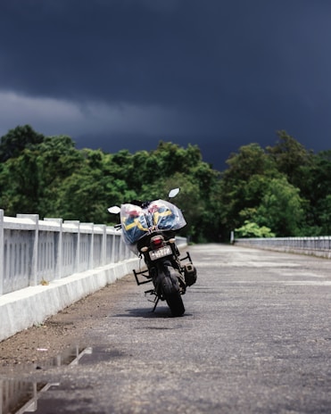 A motorcycle is parked on a deserted road with a backdrop of dark, stormy skies and lush green trees. The vehicle is positioned at an angle, showing its back end. Plastic-wrapped items are strapped to the motorcycle.