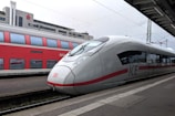 Two trains are stationed at a railway platform, with a modern white and red high-speed train in the foreground and a red double-decker train in the background. The platform is covered partially by a roof, and there are overhead electrical wires visible.