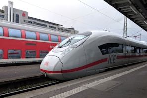 Two trains are stationed at a railway platform, with a modern white and red high-speed train in the foreground and a red double-decker train in the background. The platform is covered partially by a roof, and there are overhead electrical wires visible.
