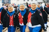 A group of people wearing colorful carnival costumes and expressive masks, featuring oversized faces with exaggerated features. They are dressed in bright red shirts, blue pants, and black jackets adorned with pins and patches. The scene is lively, taking place among a crowd of onlookers, suggesting a festive outdoor event.
