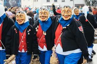 A group of people wearing colorful carnival costumes and expressive masks, featuring oversized faces with exaggerated features. They are dressed in bright red shirts, blue pants, and black jackets adorned with pins and patches. The scene is lively, taking place among a crowd of onlookers, suggesting a festive outdoor event.