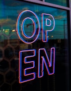 A brightly illuminated neon sign displaying the word 'OPEN' in bold blue and red colors, reflecting off a glass surface with a hexagonal pattern. The urban street scene outside is visible in the background during twilight, with a mix of ambient natural and artificial lighting.