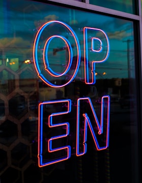 A brightly illuminated neon sign displaying the word 'OPEN' in bold blue and red colors, reflecting off a glass surface with a hexagonal pattern. The urban street scene outside is visible in the background during twilight, with a mix of ambient natural and artificial lighting.