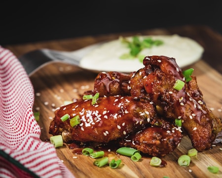 A pile of crispy chicken wings glazed with a rich, sticky sauce, garnished with sesame seeds and chopped green onions, resting on a wooden board. A red and white striped napkin is partially visible to the left, and a dish with creamy dip sits in the background, slightly out of focus.