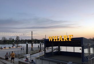 A waterfront area in the evening with a prominent sign reading 'WHARF.' String lights are draped between poles, illuminating the space as people stroll along the promenade. The background features a calm river with trees along the opposite bank, and a soft glow in the sky from the setting sun.