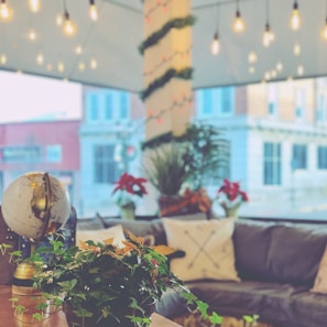 An interior space features a cozy seating arrangement with a gray couch adorned by patterned pillows. A globe and potted plants, including ivy and poinsettias, are placed on a table in the foreground. The background displays a large window offering a view of a building exterior. Warm-hued string lights hang from above, lending a festive ambiance to the setting.