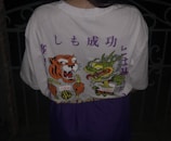 A person is wearing a white t-shirt featuring a vibrant design on the back. The design includes an illustrated tiger and dragon holding chopsticks, with bold colors and stylized elements. There are also some Japanese characters printed above the illustrations.