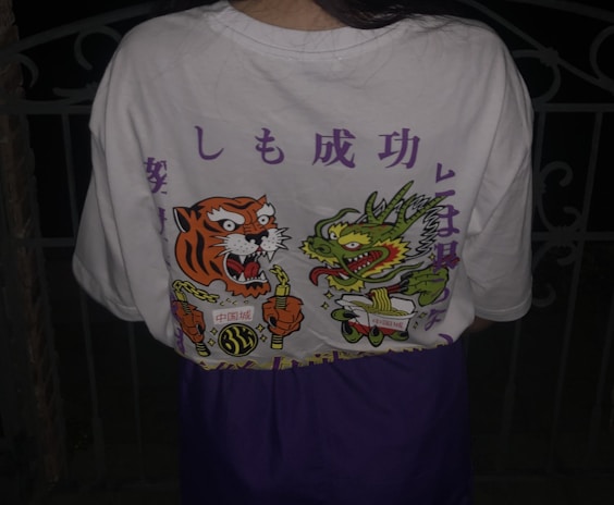 A person is wearing a white t-shirt featuring a vibrant design on the back. The design includes an illustrated tiger and dragon holding chopsticks, with bold colors and stylized elements. There are also some Japanese characters printed above the illustrations.