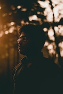 A silhouette of a person wearing glasses, set against a backdrop of trees with dappled sunlight. The warm, golden tones suggest either a sunrise or sunset, casting a peaceful and contemplative mood.
