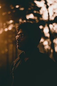 A silhouette of a person wearing glasses, set against a backdrop of trees with dappled sunlight. The warm, golden tones suggest either a sunrise or sunset, casting a peaceful and contemplative mood.