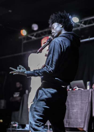 A person with an afro hairstyle is holding a microphone on stage, with a second person partly visible beside them. The setting is a dimly lit stage with colored lights overhead, and audio equipment in the background.