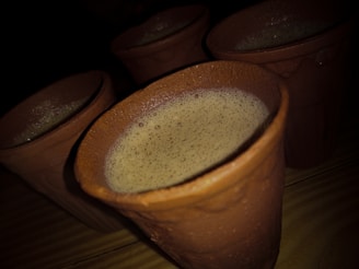Several clay mugs filled with a frothy beverage are placed on a wooden surface, with soft lighting casting shadows and highlighting the texture of the clay.