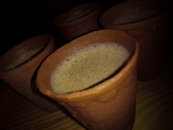 Several clay mugs filled with a frothy beverage are placed on a wooden surface, with soft lighting casting shadows and highlighting the texture of the clay.