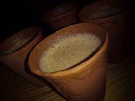 Several clay mugs filled with a frothy beverage are placed on a wooden surface, with soft lighting casting shadows and highlighting the texture of the clay.