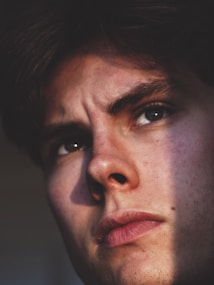A close-up portrait of a person's face, highlighting their intense expression with a focused gaze. The lighting creates dramatic shadows across their face, emphasizing the texture of their skin and the seriousness of their expression.