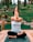 A person is practicing yoga outdoors, seated on a wooden platform with legs crossed and arms intertwined, performing a yoga pose. The background features lush greenery and trees, creating a tranquil natural setting.