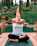 A person is practicing yoga outdoors, seated on a wooden platform with legs crossed and arms intertwined, performing a yoga pose. The background features lush greenery and trees, creating a tranquil natural setting.