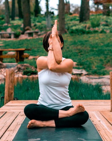 A person is practicing yoga outdoors, seated on a wooden platform with legs crossed and arms intertwined, performing a yoga pose. The background features lush greenery and trees, creating a tranquil natural setting.