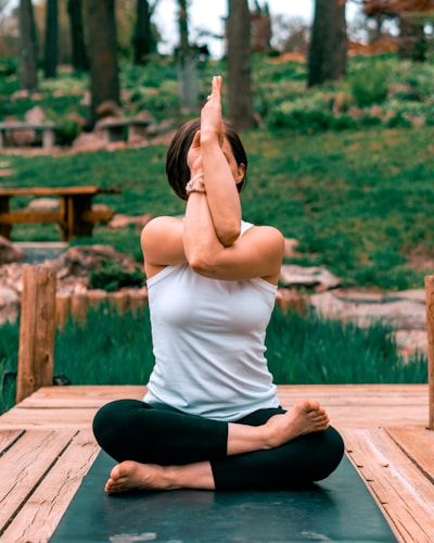 A person is practicing yoga outdoors, seated on a wooden platform with legs crossed and arms intertwined, performing a yoga pose. The background features lush greenery and trees, creating a tranquil natural setting.
