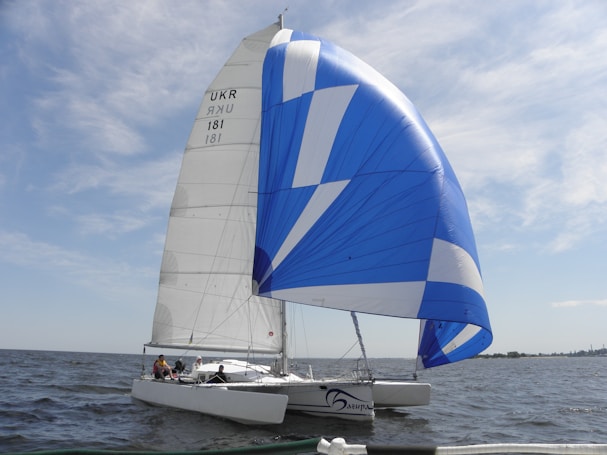 A sleek sailboat with a striking blue and white spinnaker glides smoothly over a vast body of water. The boat's hull is white, and there are several people onboard, possibly engaged in sailing activities. The sky is mostly clear with a few wispy clouds, and it appears to be a pleasant day, ideal for sailing.