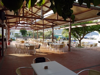 An open-air dining area with numerous empty tables and chairs is sheltered under a wooden pergola with a clear roof. The floor is tiled in a reddish-brown color and the area is surrounded by lush greenery. In the background, there are views of a marina with boats and a mountain landscape.