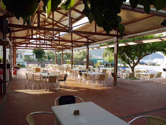 An open-air dining area with numerous empty tables and chairs is sheltered under a wooden pergola with a clear roof. The floor is tiled in a reddish-brown color and the area is surrounded by lush greenery. In the background, there are views of a marina with boats and a mountain landscape.