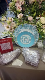A decorative setup featuring a floral arrangement with pale pink and white flowers surrounded by green foliage. In front, there are two turquoise and white geometric pattern plates, silver decorative objects resembling geological formations, a red brochure with a similar geometric pattern, and business cards on a gray tablecloth.