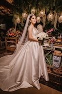 A bride stands elegantly in a satin wedding gown with a long veil, holding a bouquet of white flowers. She is surrounded by a lush backdrop of greenery, colorful flowers, and softly glowing lanterns hanging overhead. The scene is set against a rustic wooden table adorned with decorative elements.