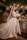 A bride stands elegantly in a satin wedding gown with a long veil, holding a bouquet of white flowers. She is surrounded by a lush backdrop of greenery, colorful flowers, and softly glowing lanterns hanging overhead. The scene is set against a rustic wooden table adorned with decorative elements.