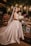 A bride stands elegantly in a satin wedding gown with a long veil, holding a bouquet of white flowers. She is surrounded by a lush backdrop of greenery, colorful flowers, and softly glowing lanterns hanging overhead. The scene is set against a rustic wooden table adorned with decorative elements.