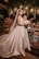A bride stands elegantly in a satin wedding gown with a long veil, holding a bouquet of white flowers. She is surrounded by a lush backdrop of greenery, colorful flowers, and softly glowing lanterns hanging overhead. The scene is set against a rustic wooden table adorned with decorative elements.