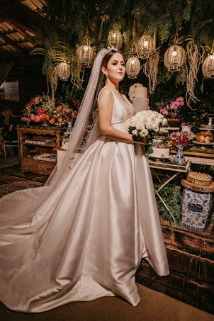 A bride stands elegantly in a satin wedding gown with a long veil, holding a bouquet of white flowers. She is surrounded by a lush backdrop of greenery, colorful flowers, and softly glowing lanterns hanging overhead. The scene is set against a rustic wooden table adorned with decorative elements.