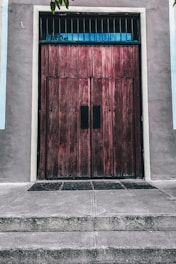 A large wooden door with a rustic, weathered texture is situated in a concrete wall. The door features vertical panels and small barred windows at the top. It is flanked by smooth, pastel-colored walls and sits above a series of concrete steps leading up to it. A few small leaves are visible in the top corners, suggesting nearby trees or plants.