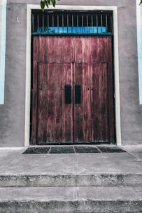A large wooden door with a rustic, weathered texture is situated in a concrete wall. The door features vertical panels and small barred windows at the top. It is flanked by smooth, pastel-colored walls and sits above a series of concrete steps leading up to it. A few small leaves are visible in the top corners, suggesting nearby trees or plants.