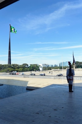 A guard in traditional uniform stands at attention beside a large plaza with a pole carrying a vibrant flag. The scene includes a modern architectural backdrop, vehicles, pedestrians, and a reflective body of water.